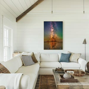 May include: A vertical wall art print featuring a night sky scene with a vibrant purple and orange Milky Way galaxy stretching above a large, illuminated rock formation on a beach, with a clear reflection visible in the wet sand below.