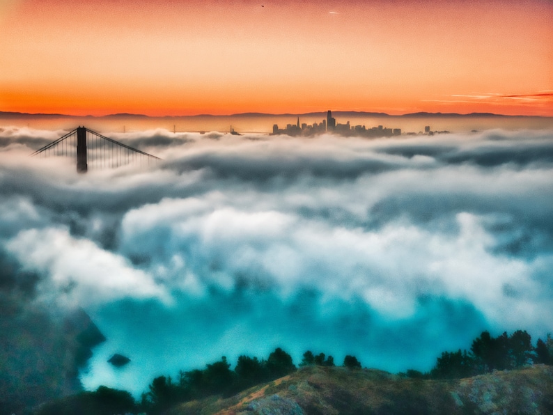 May include: A panoramic view of the Golden Gate Bridge and the San Francisco skyline emerging from a thick layer of low-hanging fog. The scene is illuminated by a vibrant orange sunset sky, contrasting with the deep blue water below the clouds.