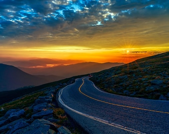 May include: A winding paved road leads up a mountainside towards a bright orange sunrise. The sky is a mix of blue and orange, with clouds casting shadows on the landscape.