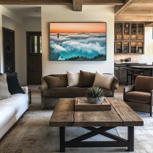 May include: A large rectangular wall art print featuring a landscape of the Golden Gate Bridge emerging from a thick layer of fog under a sunset sky with orange and blue hues, displayed in a black frame above a brown sofa in a living room.