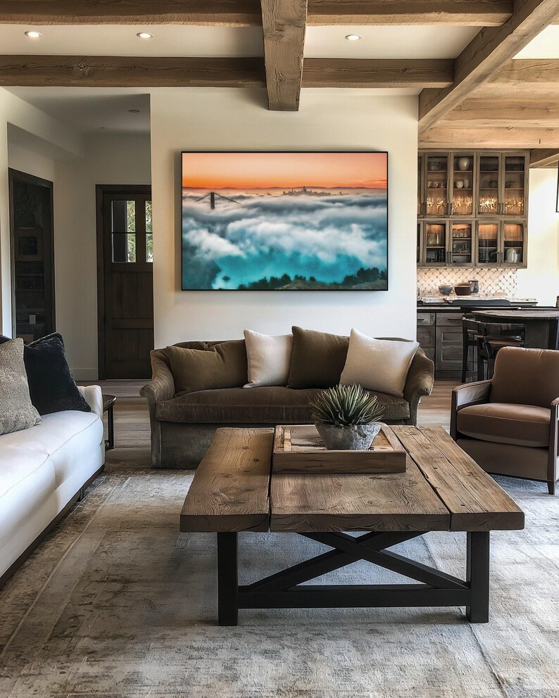 May include: A large rectangular wall art print featuring a landscape of the Golden Gate Bridge emerging from a thick layer of fog under a sunset sky with orange and blue hues, displayed in a black frame above a brown sofa in a living room.