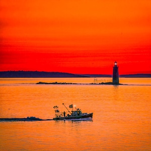 May include: A fishing boat sails across calm water under a vibrant orange and red sunset. A lighthouse stands in the distance, its light illuminated. The sky and water reflect the warm colors of the setting sun.