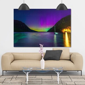 May include: A large rectangular canvas print depicting a vibrant night scene. The image showcases a body of water with a sailboat, mountains, and a colorful aurora borealis display in shades of purple, green, and yellow. The print is displayed above a beige sofa.