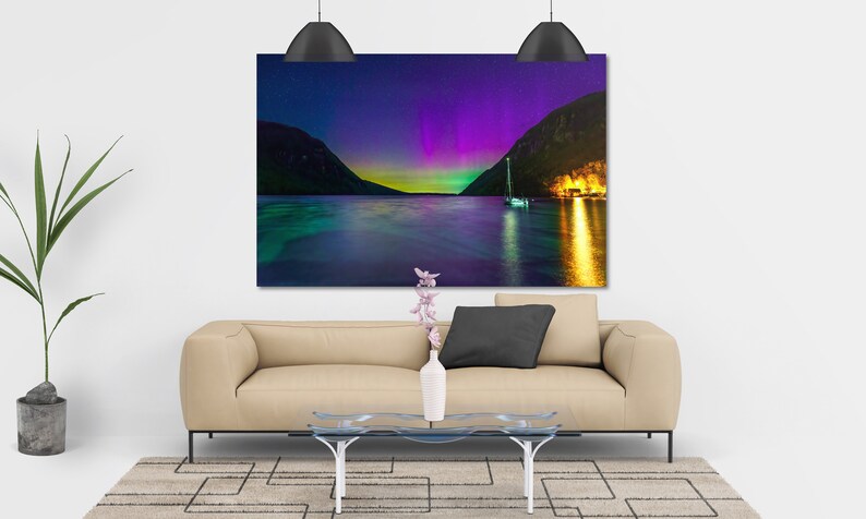 May include: A large rectangular canvas print depicting a vibrant night scene. The image showcases a body of water with a sailboat, mountains, and a colorful aurora borealis display in shades of purple, green, and yellow. The print is displayed above a beige sofa.