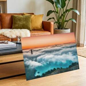 May include: A rectangular wall art print featuring a landscape photograph of the Golden Gate Bridge partially obscured by a thick layer of low-hanging fog, with a vibrant orange sunset sky above and dark green hills visible in the foreground.