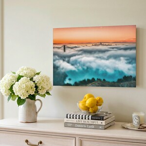 May include: A rectangular wall art print featuring a landscape photograph of the Golden Gate Bridge partially obscured by thick fog under a sunset sky with orange and blue hues, displayed in a room setting with a vase of white hydrangeas.