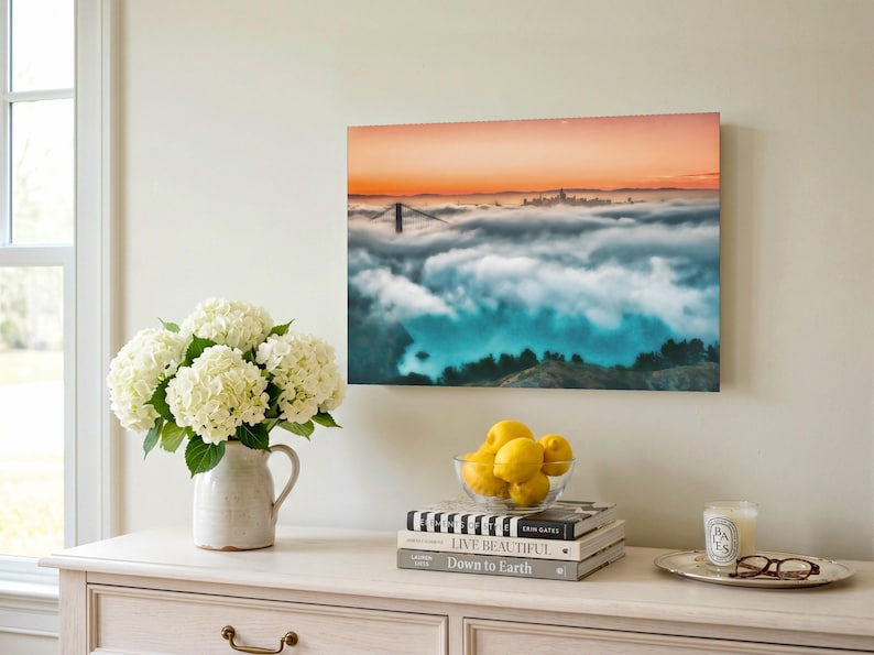 May include: A rectangular wall art print featuring a landscape photograph of the Golden Gate Bridge partially obscured by thick fog under a sunset sky with orange and blue hues, displayed in a room setting with a vase of white hydrangeas.