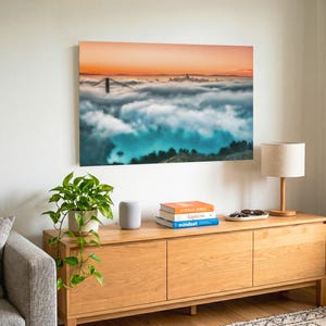 May include: A rectangular wall art print featuring a landscape photograph of the Golden Gate Bridge partially obscured by thick fog under a vibrant orange sunset sky, displayed in a room with a wooden console table and a potted plant.