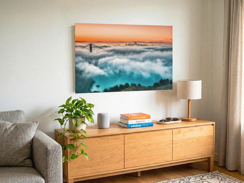 May include: A rectangular wall art print featuring a landscape photograph of the Golden Gate Bridge partially obscured by thick fog under a vibrant orange sunset sky, displayed in a room with a wooden console table and a potted plant.