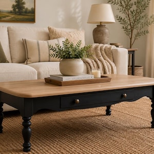 May include: A rectangular coffee table with a light wood top and black base. The table features a single drawer with brass knobs and turned legs. A tray with books and a vase of greenery sits on top, with a neutral-toned sofa in the background.