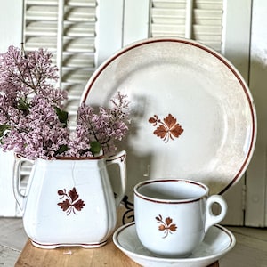 May include: A white ceramic pitcher, plate, cup, and saucer set with a brown leaf design. The pitcher holds purple flowers. The plate and cup have a brown rim. The set is on a wooden tray.