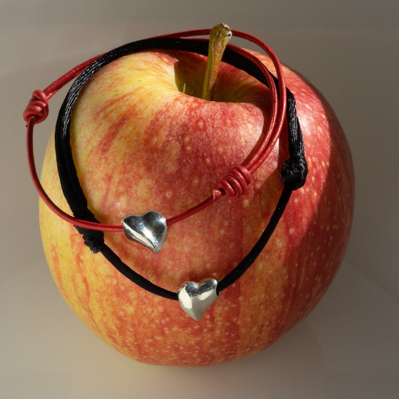 May include: A red leather bracelet with a black leather bracelet wrapped around it. Both bracelets have silver heart charms. The bracelets are wrapped around a red apple.