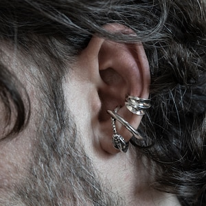 May include: A person's ear with three silver ear cuffs. The ear cuffs are stacked on top of each other. The top ear cuff is a simple band, the middle ear cuff is a curved band, and the bottom ear cuff is a more intricate design.