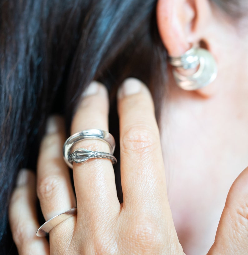 May include: A close-up of a hand wearing three silver rings. The rings are stacked on the middle finger. The top ring is a simple band, the middle ring is a textured band, and the bottom ring has a decorative design.