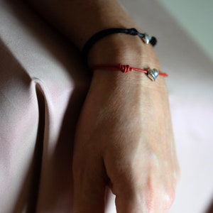 May include: A close-up of a person's wrist wearing two bracelets. The top bracelet is black with a silver charm. The bottom bracelet is red with a silver heart charm.