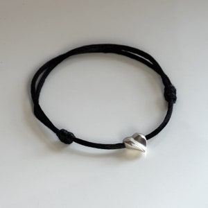 May include: A black cord bracelet with a silver heart charm. The heart charm is a simple, modern design.