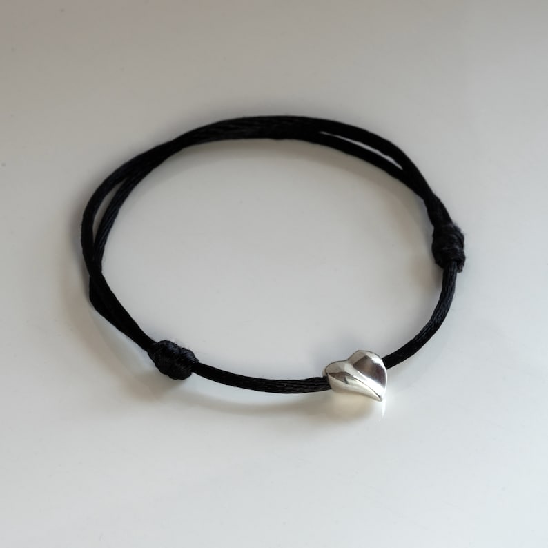 May include: A black cord bracelet with a silver heart charm. The heart charm is a simple, modern design.