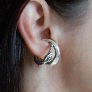 May include: Silver snake ear cuff with a coiled design. The cuff is worn on the upper part of the ear.