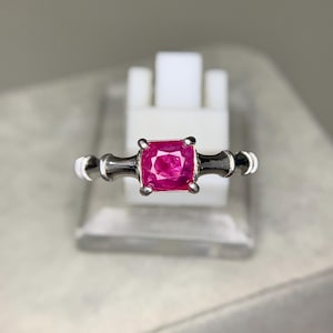 May include: A silver ring featuring a square, vibrant pink gemstone. The ring's band has a unique design with a dark gray center and white accents. The gemstone is the focal point, set in a classic four-prong setting.