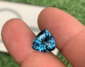Puede incluir: Una piedra preciosa azul de corte trillion con una superficie facetada que refleja la luz. La piedra muestra un color azul profundo y vibrante y una forma triangular, con cortes geométricos precisos que crean un aspecto brillante.