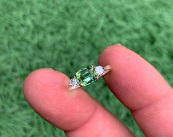 Puede incluir: Un anillo de plata con una piedra preciosa verde rectangular vibrante como pieza central, flanqueada por dos piedras blancas más pequeñas en forma de pera. El anillo se encuentra sobre un fondo de hierba verde.