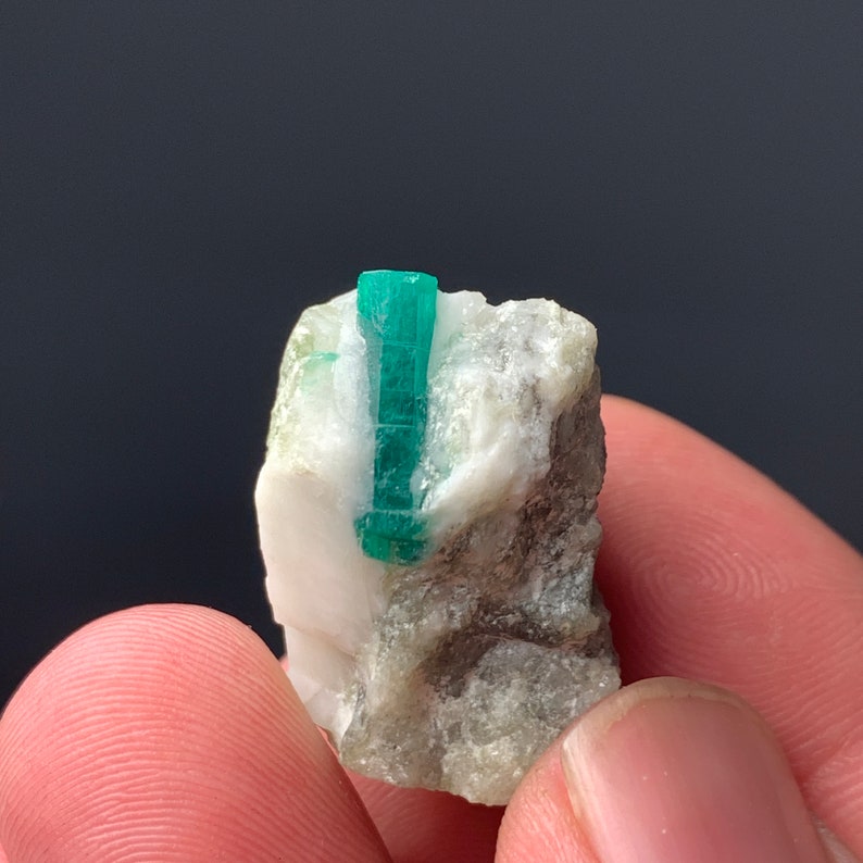Puede incluir: Un peque&ntilde;o cristal de esmeralda verde incrustado en una matriz de roca blanca y gris. El cristal mide aproximadamente 2,5 cm de largo y tiene una punta afilada.