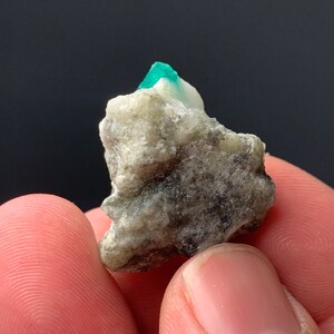 Puede incluir: Una peque&ntilde;a roca gris rugosa con un cristal de esmeralda verde brillante en la parte superior.