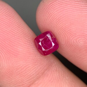 May include: A deep red gemstone with a square cushion cut. The gemstone is faceted and has a high polish.