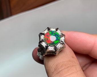 May include: A silver ring with a large, round opal gemstone. The opal showcases a vibrant play of colour, with red, green, and white hues. The ring's setting has a unique, spiky design, adding to its distinctive appearance.