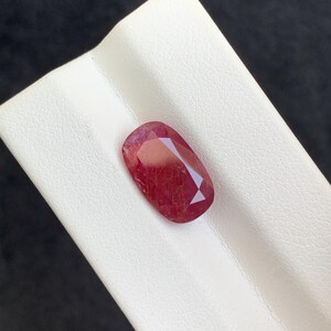 May include: A faceted, oval-shaped ruby gemstone with a deep red hue. The gem is set against a white surface, showcasing its polished facets and internal clarity. The ruby's color ranges from a vibrant red to a slightly lighter shade in the center.