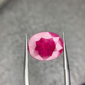 Pode incluir: Uma gema oval de cor rosa vibrante com corte facetado, segurada por pinças sobre um fundo cinza.