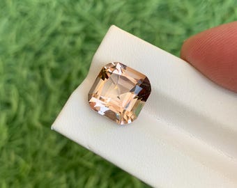 Puede incluir: Una gema facetada, de corte cuadrado, con un degradado de color melocotón y marrón. La gema está colocada sobre una superficie blanca, con un fondo verde borroso. Las facetas de la gema reflejan la luz.