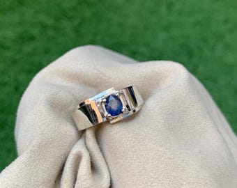 Puede incluir: Un anillo de plata con una gran gema azul de forma ovalada. El anillo tiene un diseño moderno con una banda ancha y detalles geométricos. La gema está engastada en un bisel.