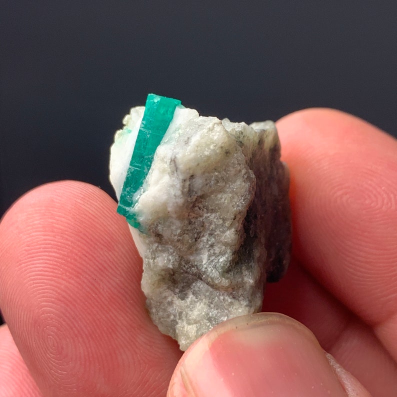 Puede incluir: Un peque&ntilde;o cristal de esmeralda verde incrustado en una matriz blanca, gris y negra. El cristal mide aproximadamente 2,5 cm de largo y tiene una forma puntiaguda.