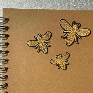 Peut inclure: Trois écussons dorés en forme d'abeilles avec des contours noirs sont disposés sur un cahier à spirales marron. Les abeilles ont des ailes et des corps détaillés. La reliure à spirale du cahier est argentée.