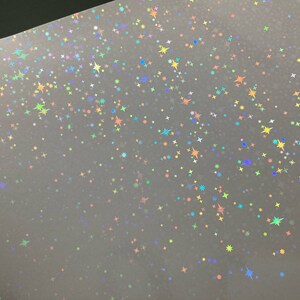 May include: A sheet of iridescent holographic paper with a scattered pattern of small stars in various colors, including blue, green, yellow, and pink.
