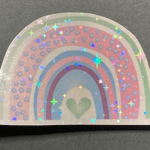 May include: A holographic rainbow sticker with a green heart in the center. The rainbow is surrounded by a scattering of iridescent stars.