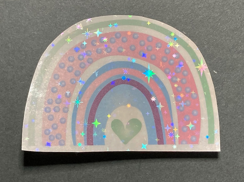 May include: A holographic rainbow sticker with a green heart in the center. The rainbow is surrounded by a scattering of iridescent stars.