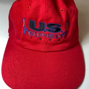 Op de afbeelding: Rode baseballpet met de geborduurde tekst "U.S. Open 1996" in zwart, blauw en goud.
