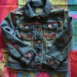 May include: A blue denim jacket with colourful embroidered trim. The jacket has a button closure and five pockets. The label on the jacket reads "Happy Little Market".