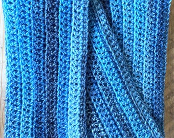 Puede incluir: Una bufanda de ganchillo rectangular con textura acanalada y una paleta de colores variados en hilos azul marino, azul real y azul cielo. La pieza está confeccionada con puntos verticales que crean un patrón consistente y elevado.