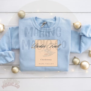 May include: A pale blue sweatshirt featuring a wine label design. The label says "Parker Knoll 1993 Chardonnay Napa Valley". The words "Making Magic Co" are faintly visible. Gold baubles are scattered around the sweatshirt.