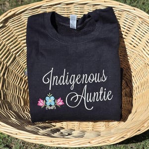 May include: Black sweatshirt with the text "Indigenous Auntie" in white cursive lettering and a small embroidered floral design in pink, blue, and green.
