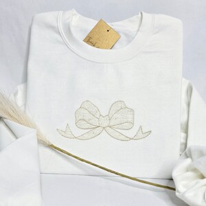 May include: White sweatshirt with a gold embroidered bow on the front. The sweatshirt has a crew neck and long sleeves.