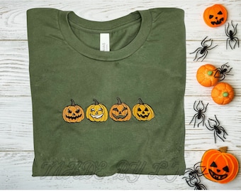 May include: A green t-shirt with a row of four embroidered orange pumpkins with different facial expressions. The pumpkins are all the same size and are arranged in a line across the center of the shirt.