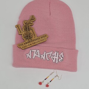 May include: A pink beanie with white text and a wooden emblem. The beanie has the text "ATA ATHS" in white. The wooden emblem has the text "THE EMBROIDERED MOM CUSTOM EMBROIDERY". A pair of earrings with red rose charms are also in the image.