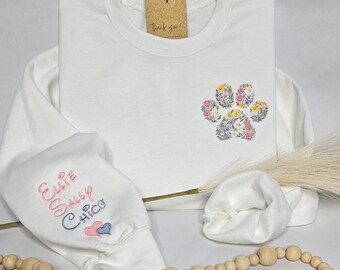 May include: A white sweatshirt with a floral paw print design. The paw print is made of embroidered flowers in shades of pink, yellow, and purple. The left sleeve has embroidered names and two hearts. A tag reads "Thank you!"