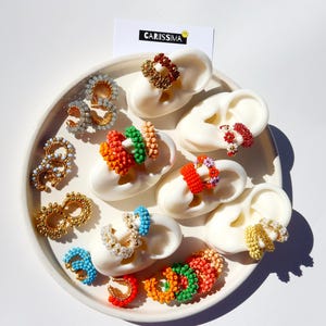 May include: A collection of colorful beaded earrings displayed on a white plate. The earrings feature various designs and colors, including gold, blue, red, orange, and green. A card with the text "CARISSIMA" is also visible.