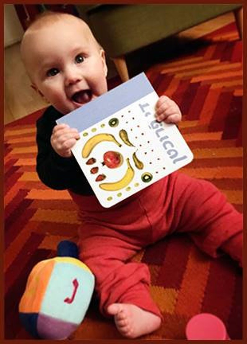 May include: A baby in a red and black outfit holds a book titled "It's Logical!" The book has a colorful design with fruit and vegetables. The baby is sitting on a red and white patterned rug.