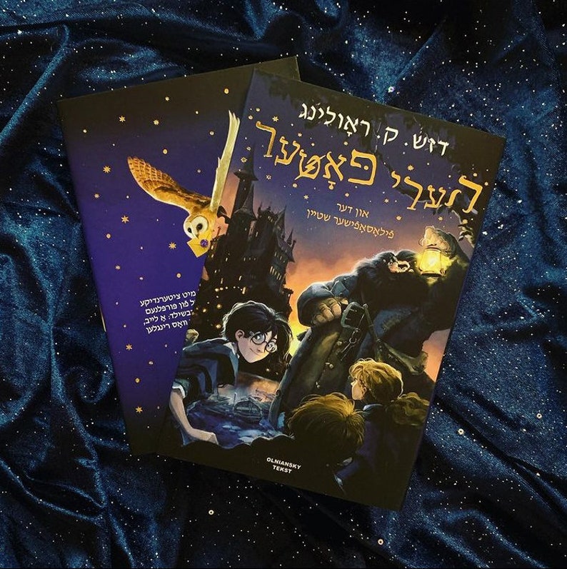 May include: A blue and purple book cover featuring a dark castle and a flying owl. The title of the book is "Harry Potter and the Philosopher's Stone" in Hebrew. The book is on a blue sparkly background.